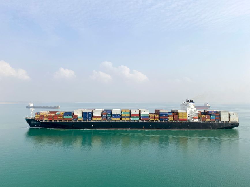 Aerial view of a large cargo ship filled with containers sailing on a calm sea under a clear sky.