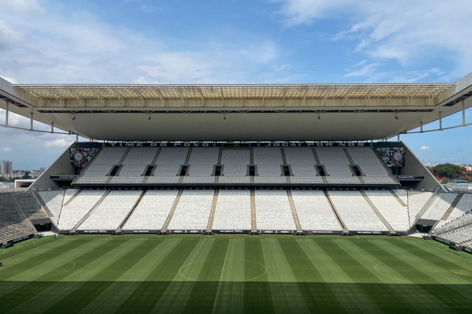 Vista ampla de uma arena de futebol vazia em São Paulo, com um campo verde sob um céu ensolarado.