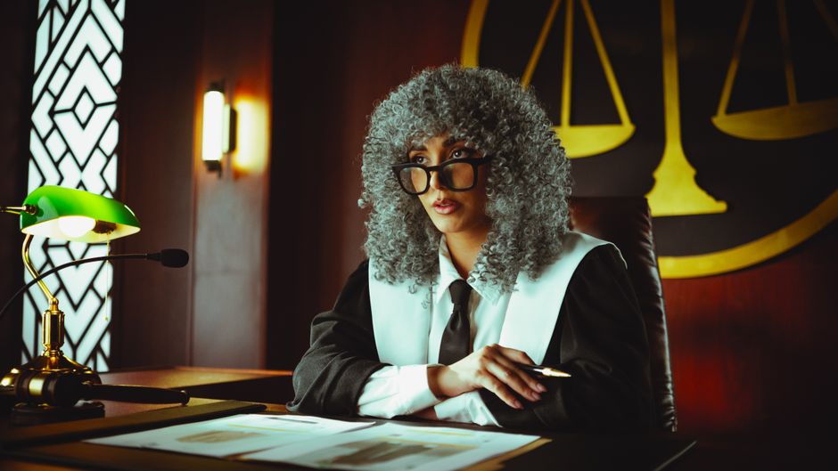 Female judge in courtroom setting, sitting at desk with justice scales in background.