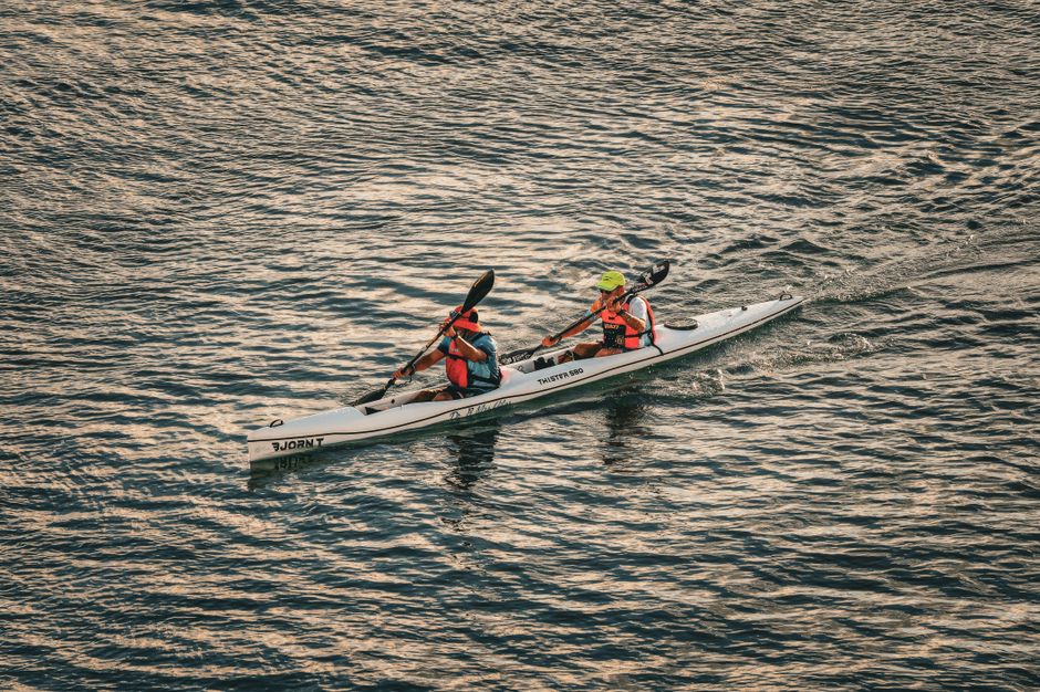 Two adults kayaking on a calm body of water, wearing life vests and helmets during the day.