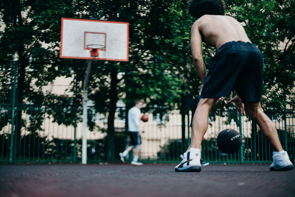 Two athletes playing basketball shirtless on an outdoor court with trees surrounding.