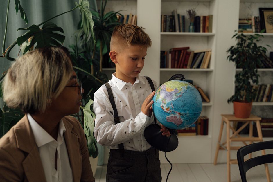 A young boy studying geography indoors with a globe and teacher nearby.