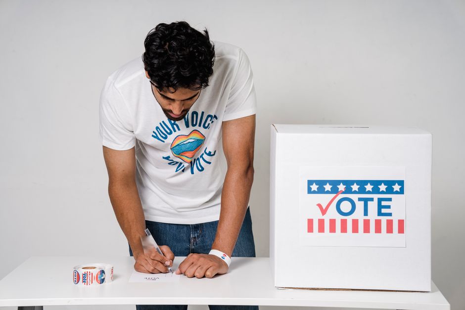 A man writes on a paper to cast his vote next to a ballot box in a studio setting.