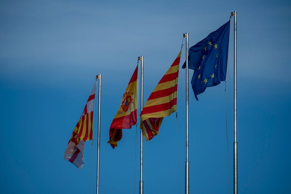 Flags of Spain, Catalonia, and Europe on flagpoles against a blue sky in Barcelona.