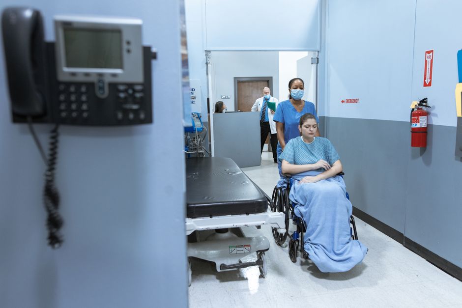 A nurse helps a patient in a wheelchair down a hospital corridor, reflecting care and medical professionalism.