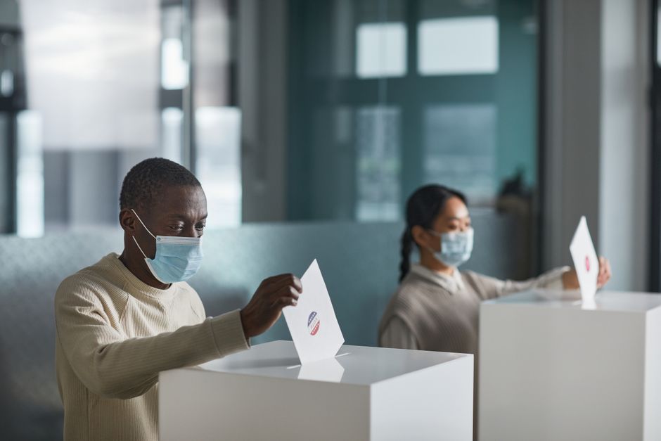 People wearing face masks cast ballots in a public indoor polling station with social distancing.