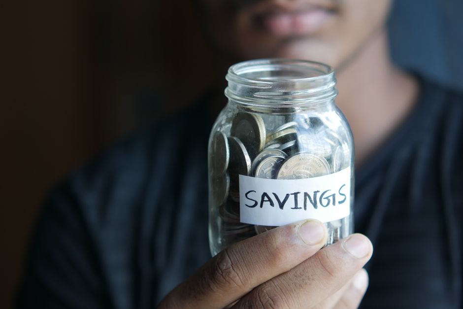 A close-up image of a person's hand holding a jar full of coins labeled 'Savings'.